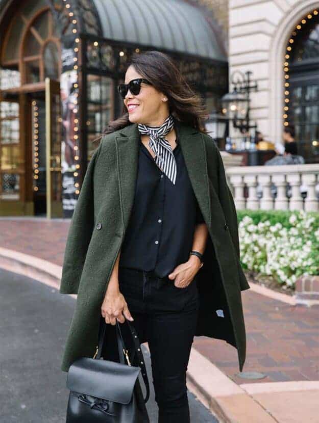 Green jacket with black jeans and black top with scarf standing in front of hotel