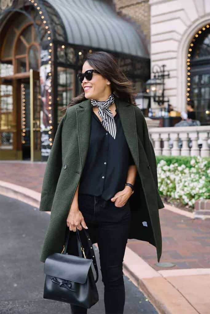 Green jacket with black jeans and black top with scarf standing in front of hotel