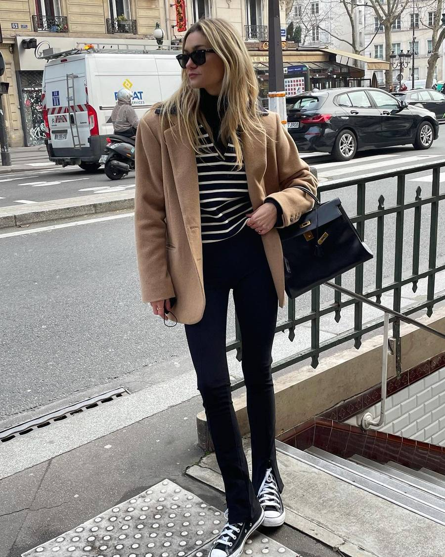 Lady wearing camel coat, striped top, black leather bag, black pants, converse high tops in Paris