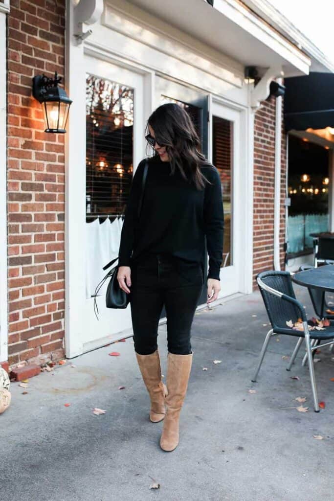 Black turtleneck and black jeans with brown tall boots and black bag standing outside a cafe.