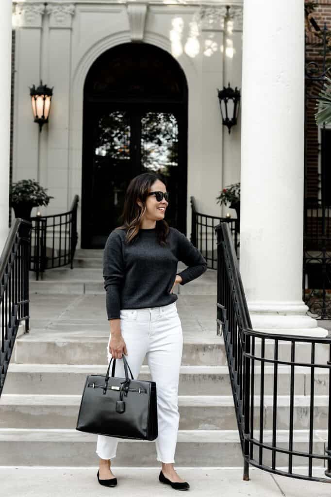 standing in front of building with grey sweater, white jeans, and black bag