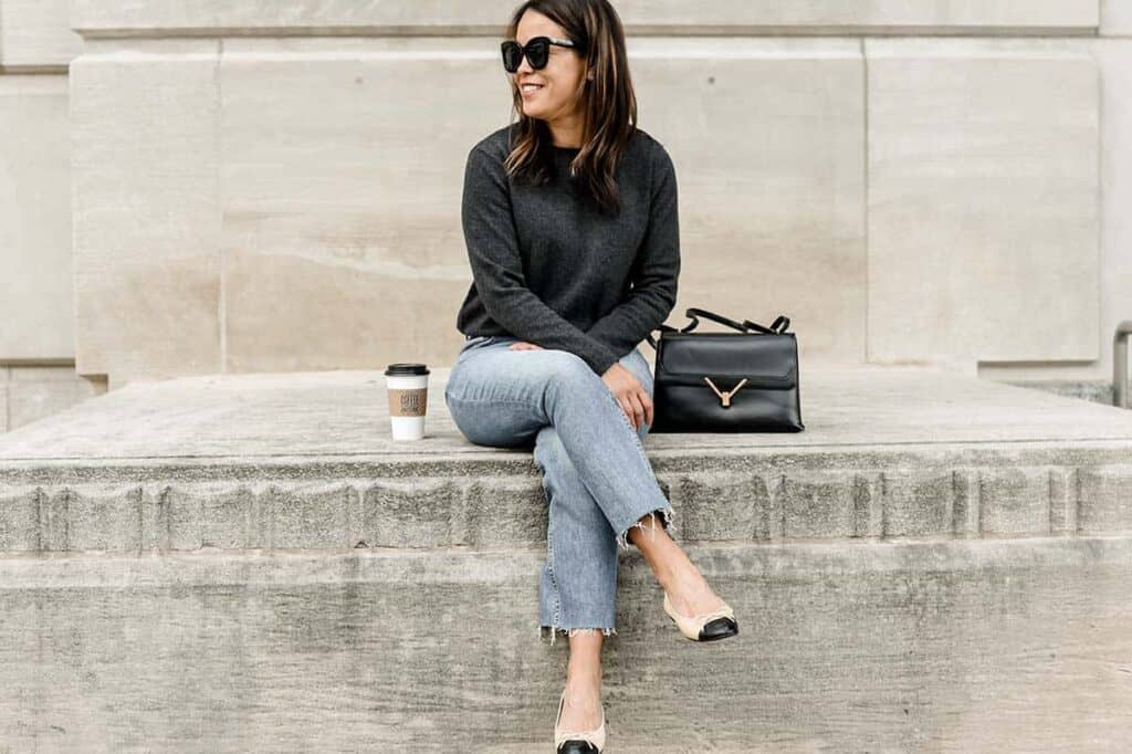 Sitting wearing dark grey cashmere sweater with sunglasses, blue denim, and flats