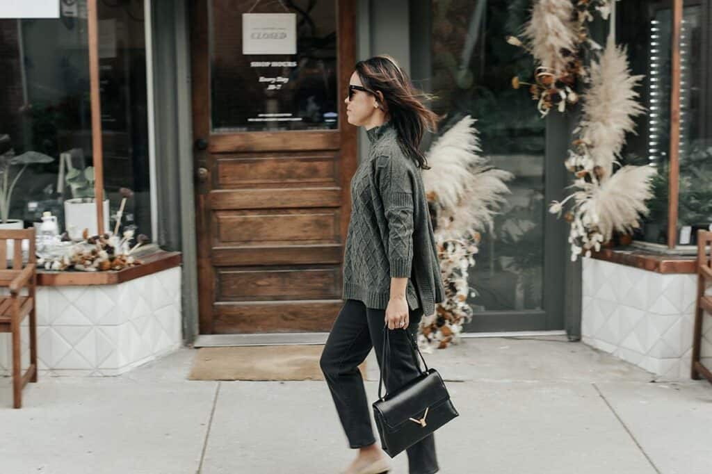 walking in a green sweater with black jeans and holding a black handbag