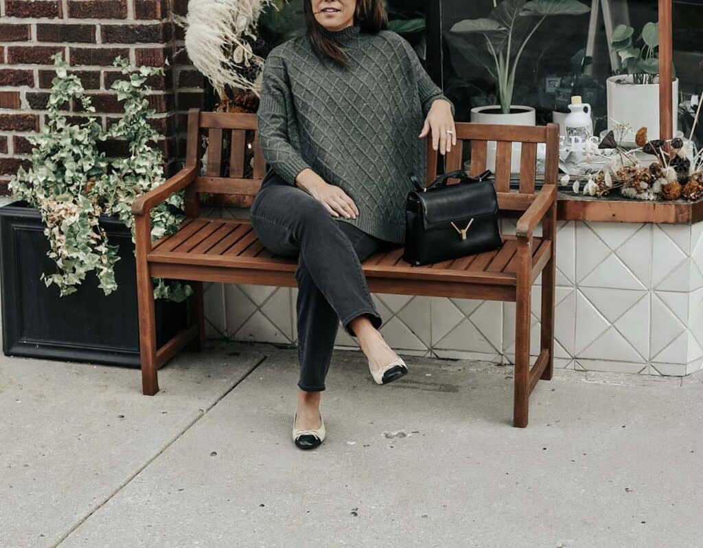 Sitting on a bench in a green mer sea sweater with flats, black jeans, and sunglasses