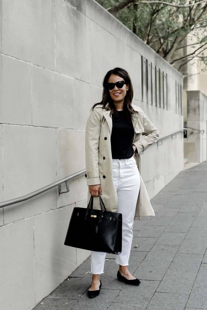 wearing a trench coat with white jeans, holding black purse, and wearing black flats 