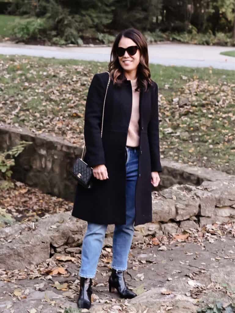 Standing in black coat with jeans, brown sweater and fall leaves in background 