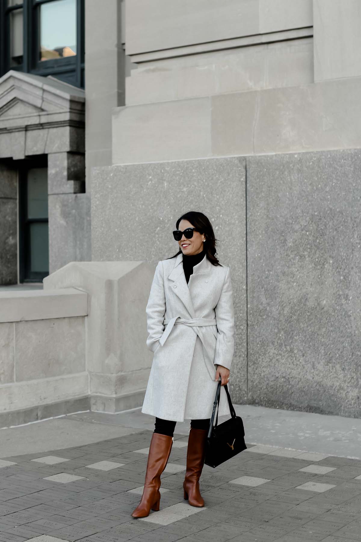 Standing in front of a building with grey coat and black bag 