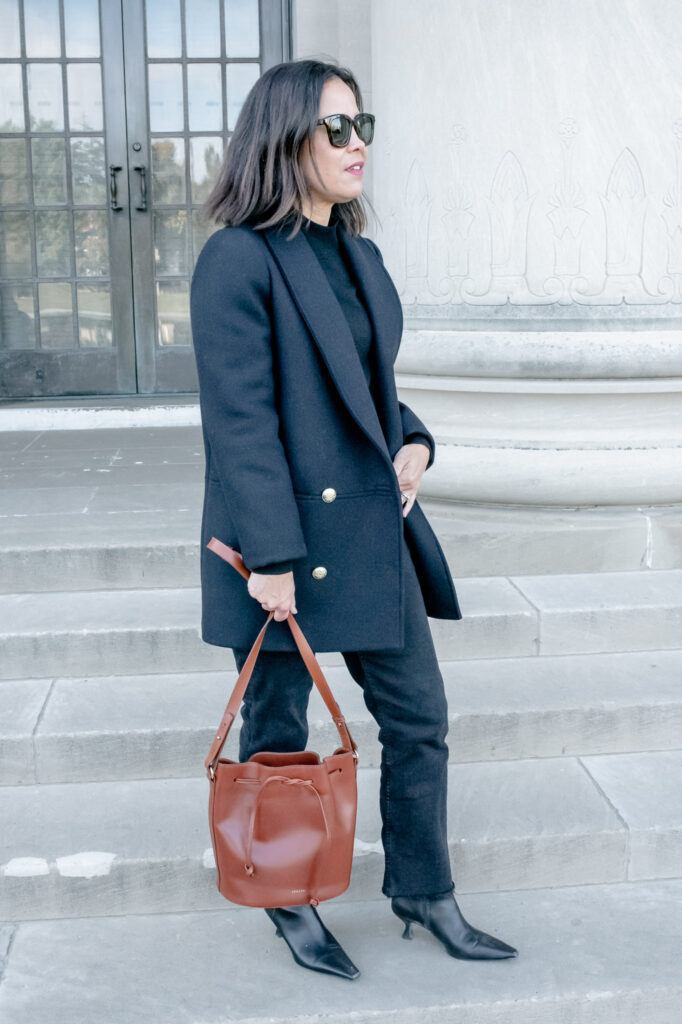 Lady wearing navy coat with black jeans, brown handbag standing against a wall