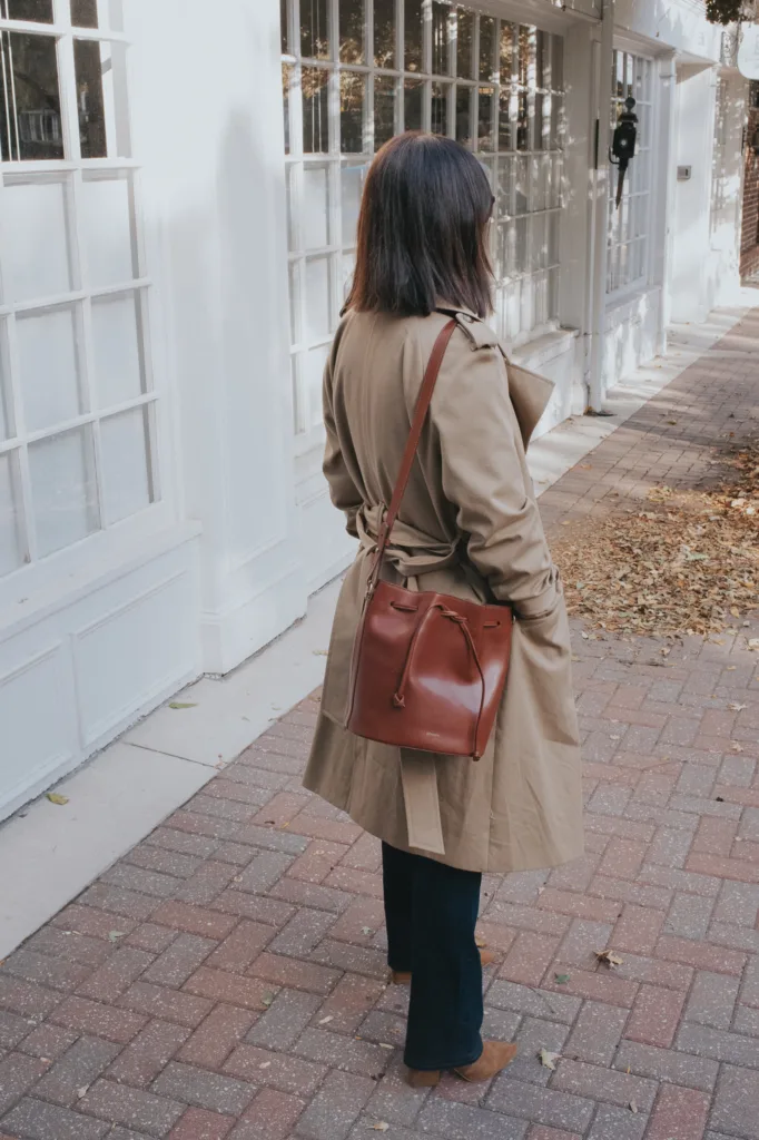 Woman standing wearing trench coat and leather bucket bag with jeans.