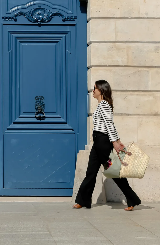 Lady wearing black pants, striped top and carrying market tote in front of blue door in Paris.