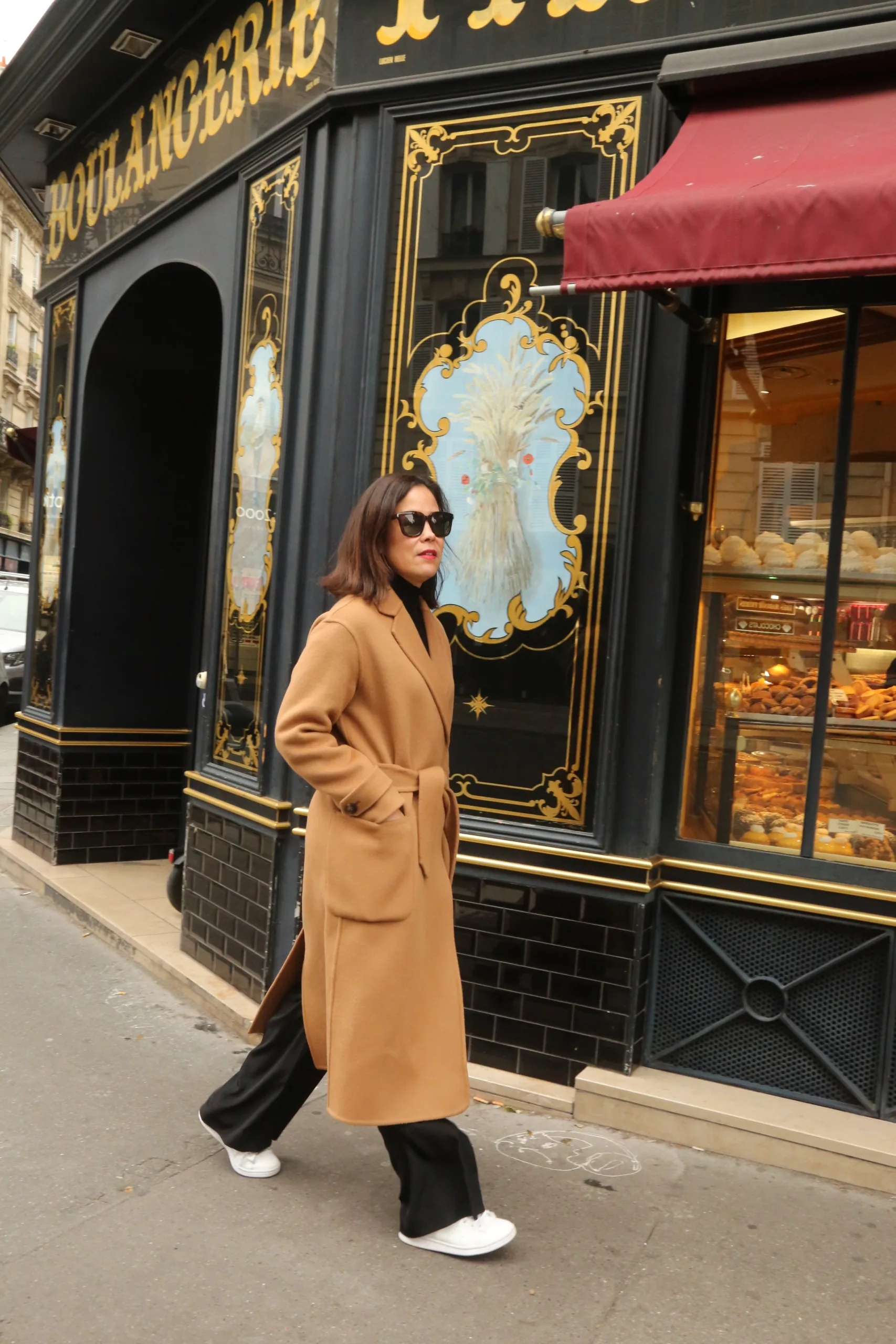 Lady wearing camel coat walking in front of a boulangerie in Paris.