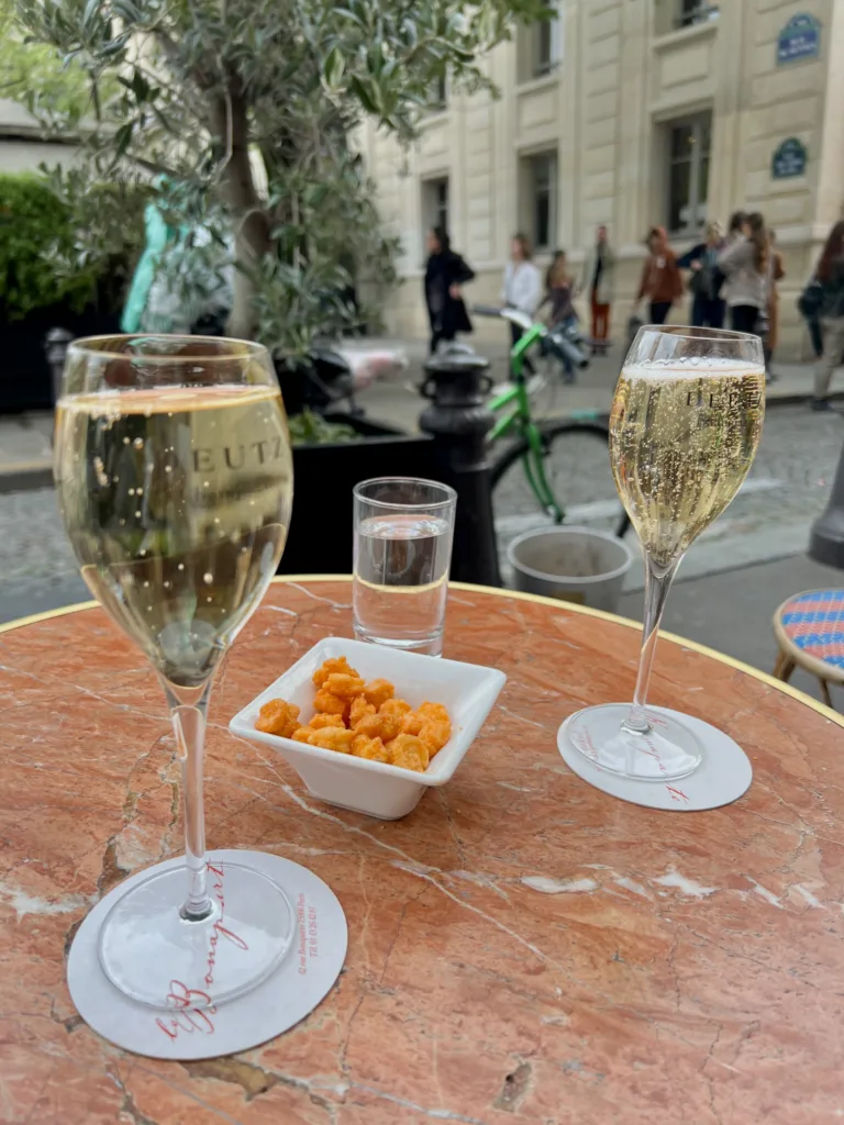 Two champagne glasses sitting on a table in Paris at a cafe with people walking in the background in saint germain des pres.