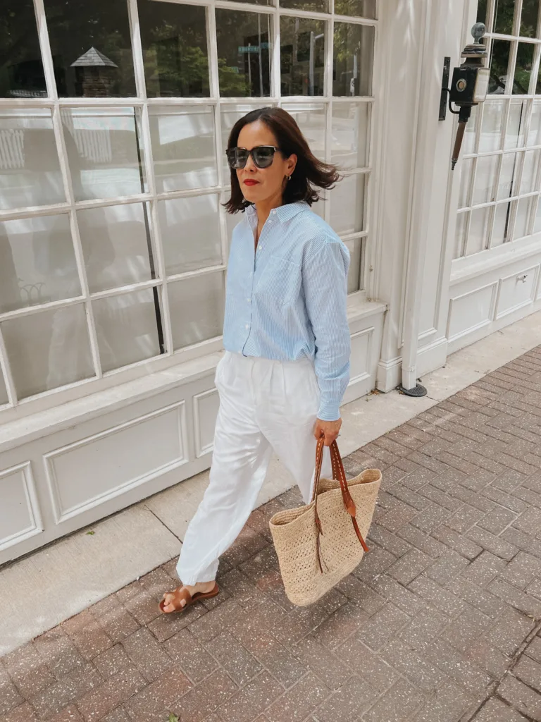 white linen pants outfit Woman walking with white linen pants, striped top, sandals, and summer tote.