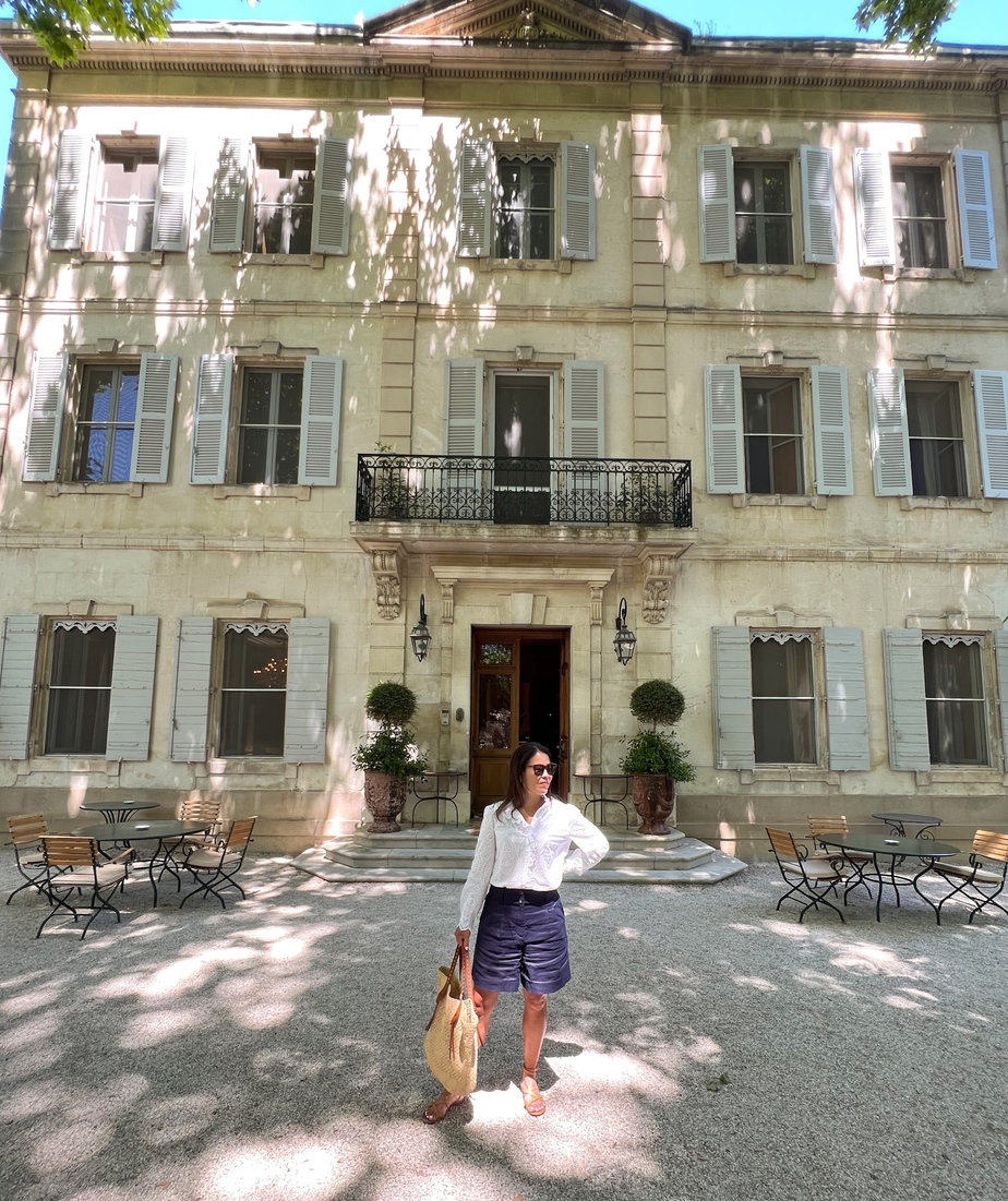 white eyelet Sézane blouse with navy linen shorts and sandals standing against a pole 