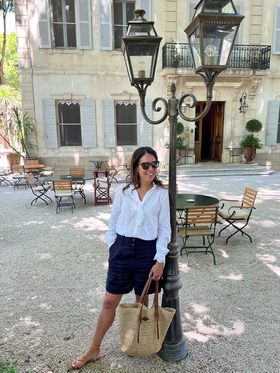 white eyelet Sézane blouse with navy linen shorts and sandals standing against a pole