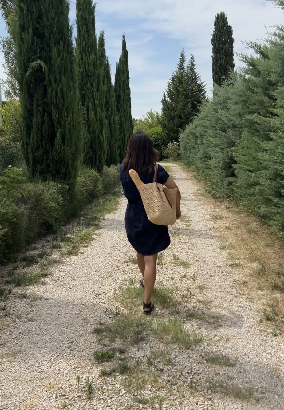 walking down gravel road wearing linen dress, sandals, carrying market bag with baguette 