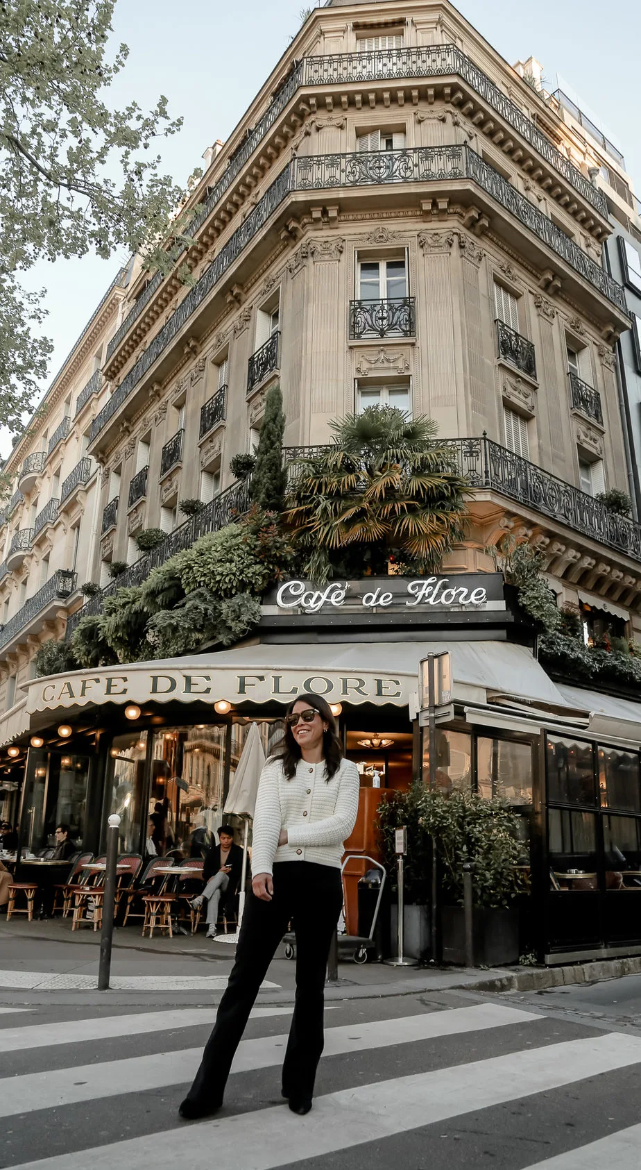 Wearing a white cardigan with black jeans and boots standing outside Cafe de Flore in Paris