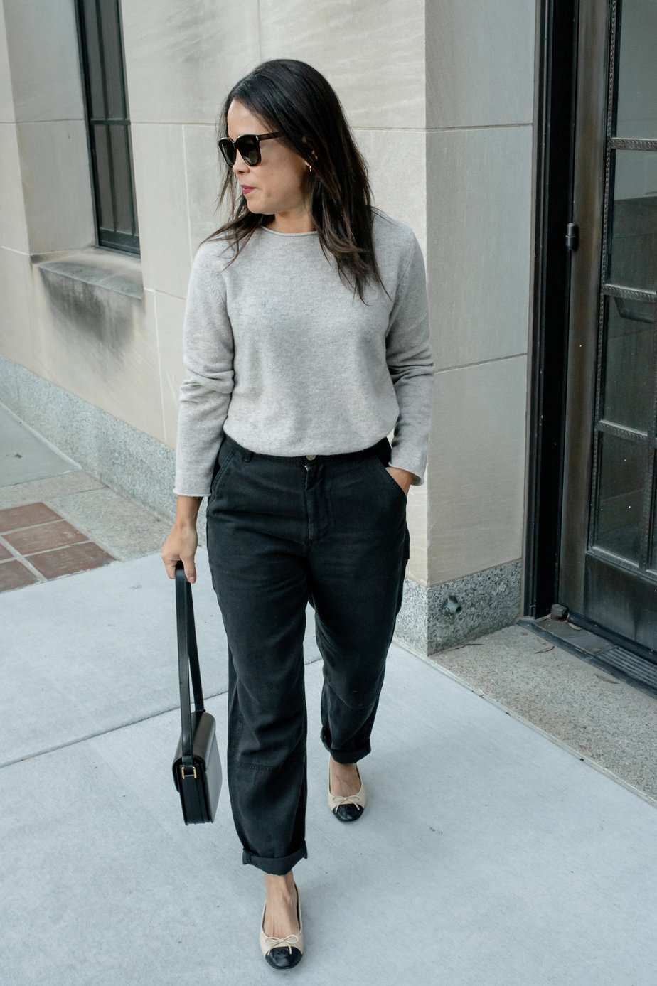 woman wearing taupe sweater with black pants, black leather bag, and chanel flats walking
