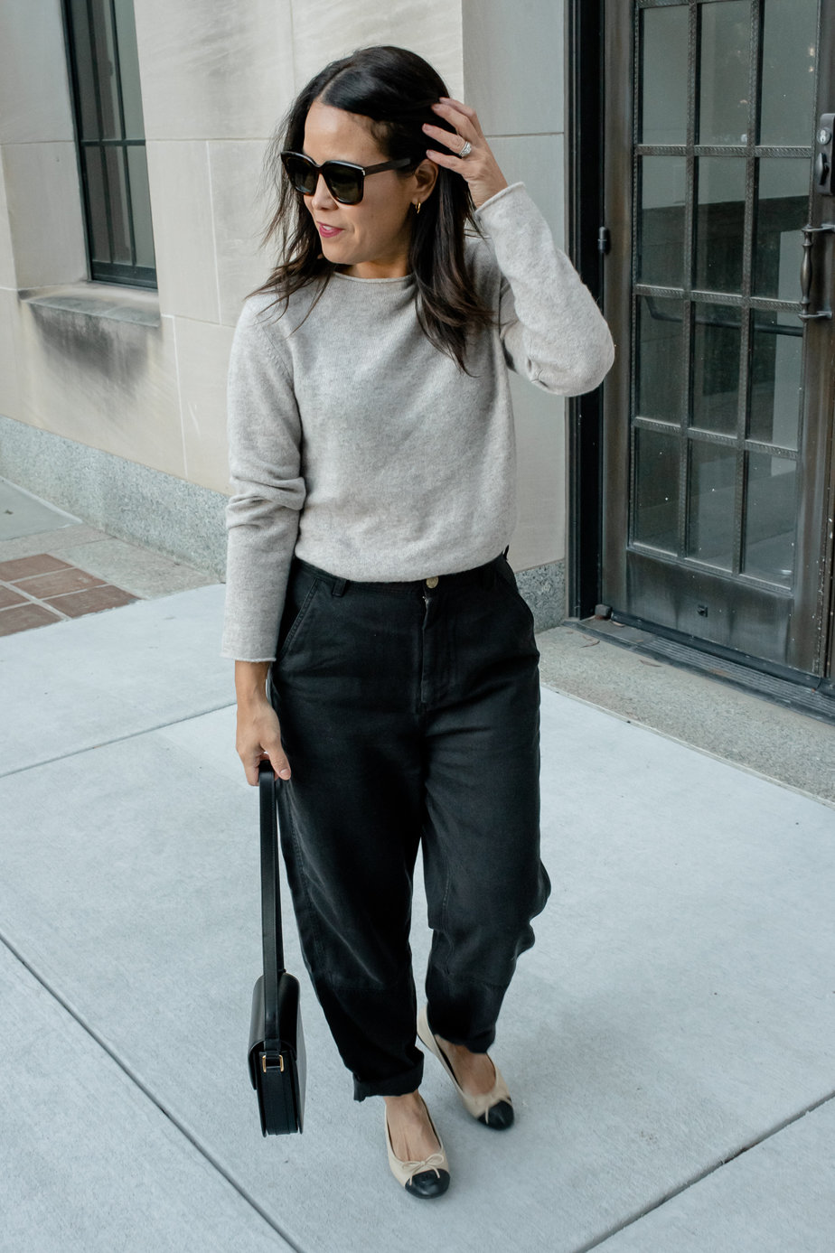 woman wearing taupe sweater with black pants, black leather bag, and chanel flats walking down the road