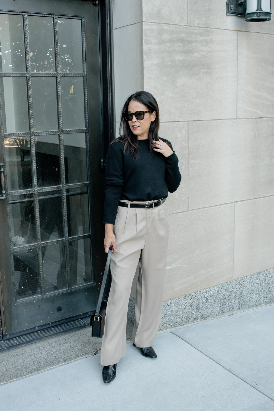 Woman wearing black sweater wearing trousers, black belt, black boots, and black leather handbag standing against a wall