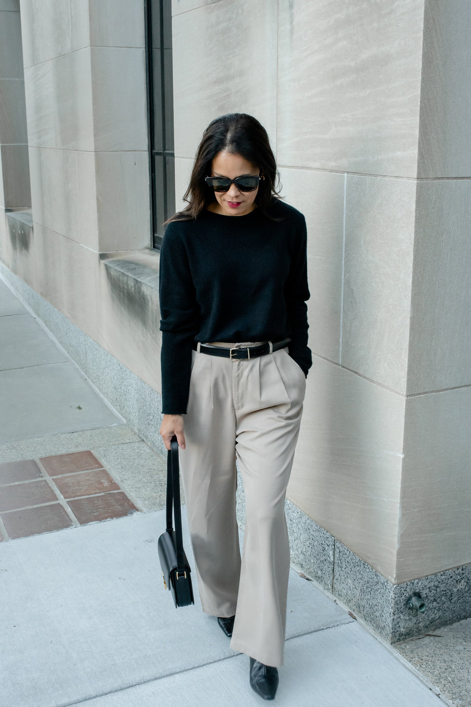 Woman wearing black sweater wearing trousers, black belt, black boots, and black leather handbag walking down the road