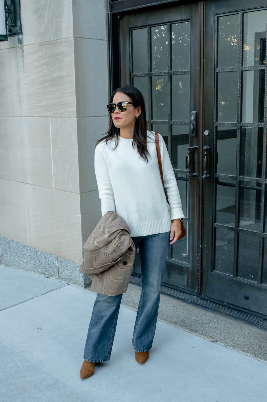 woman in cream mer sea cashmere crew sweater holding a jacket and wearing denim standing in front of a black door