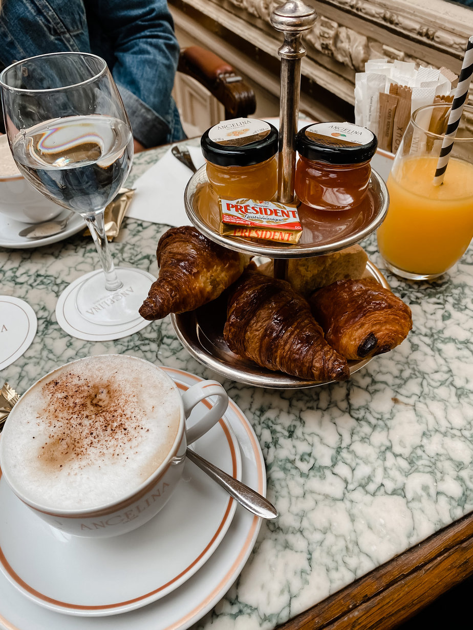 Breakfast of croissants, jam in little bottles, and packet of butter sitting in countertop tray with latte on table at Angelina's in Paris for French Fridays