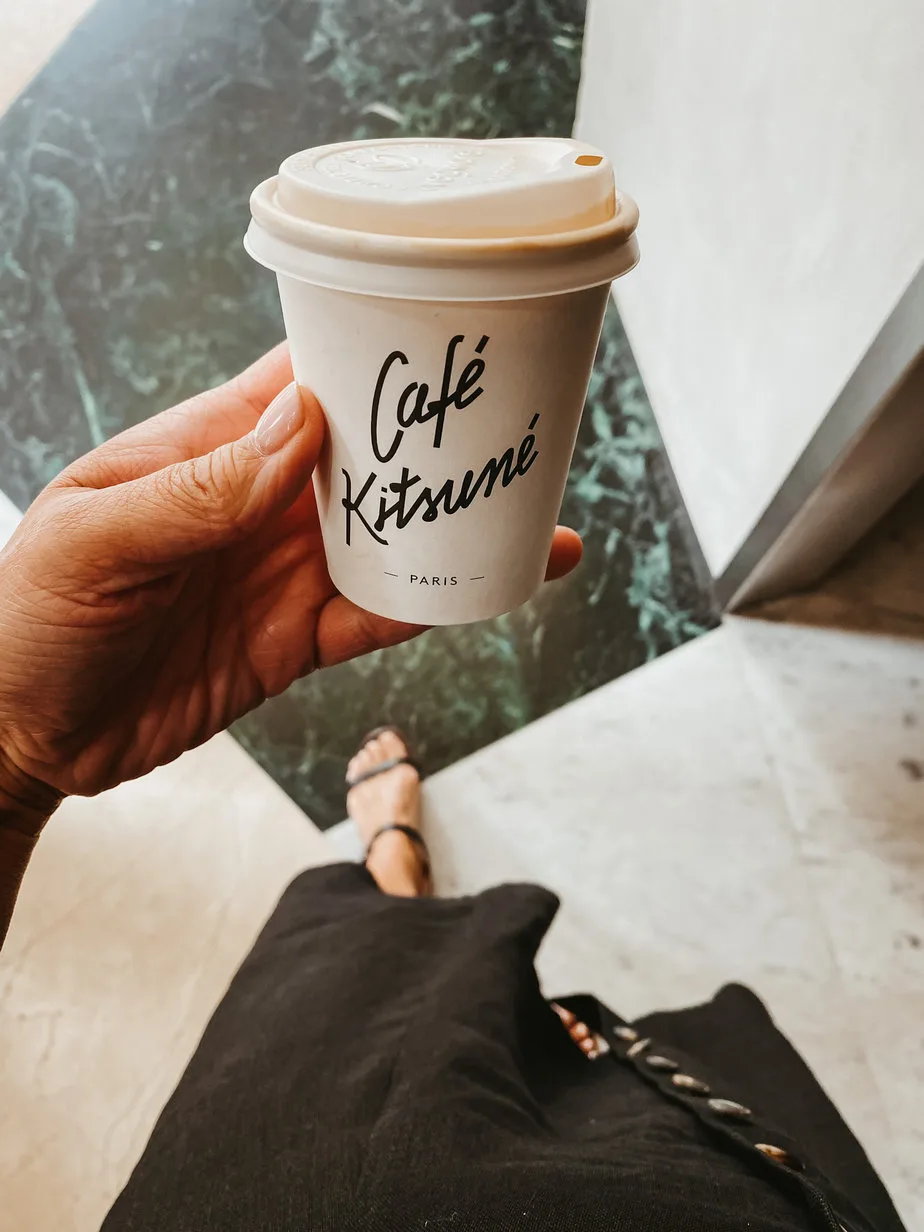 Holding a Cafe Kitsuné paper white cup with black logo in front of a black and white checkerboard floor.