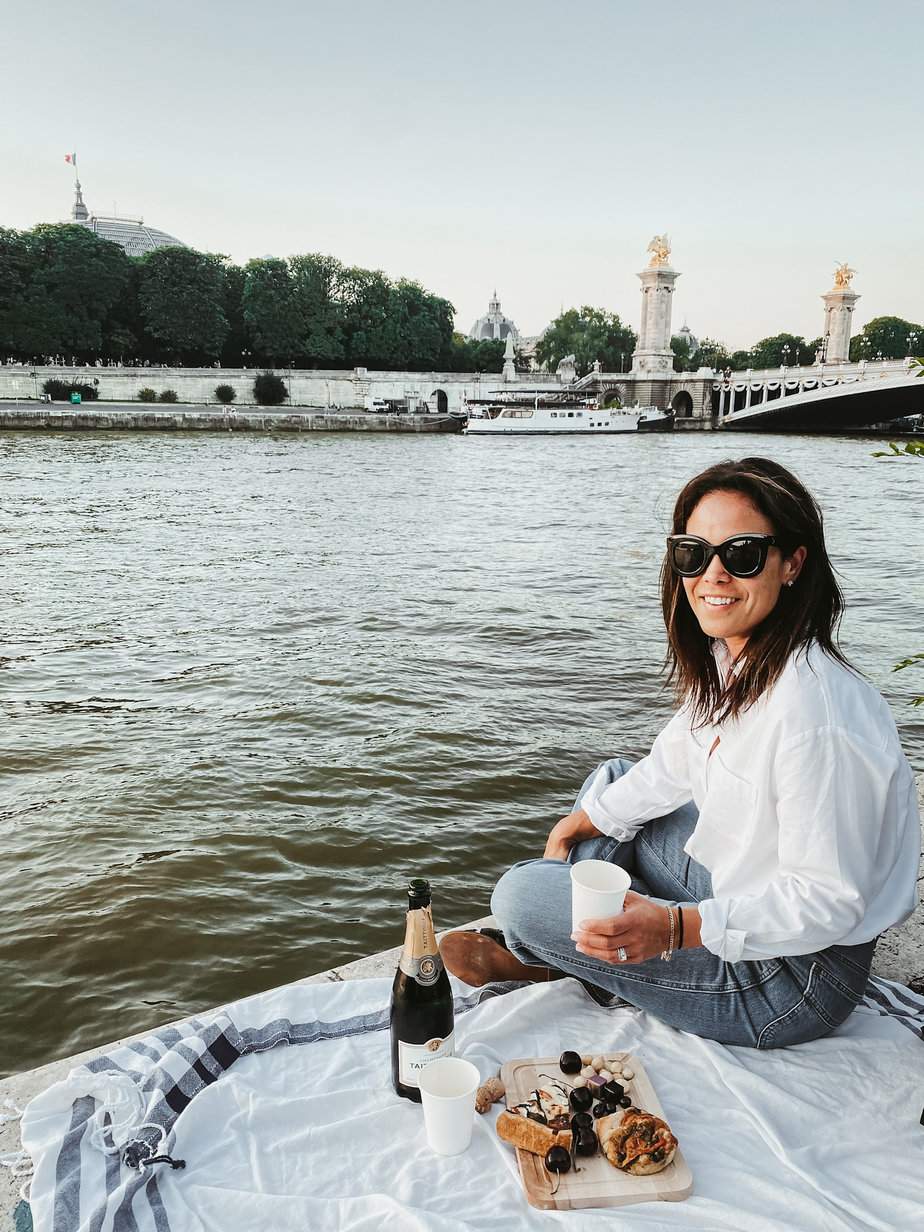 Picnic on the seine wearing a white button down and blue jeans with the point alexandre bridge in the background