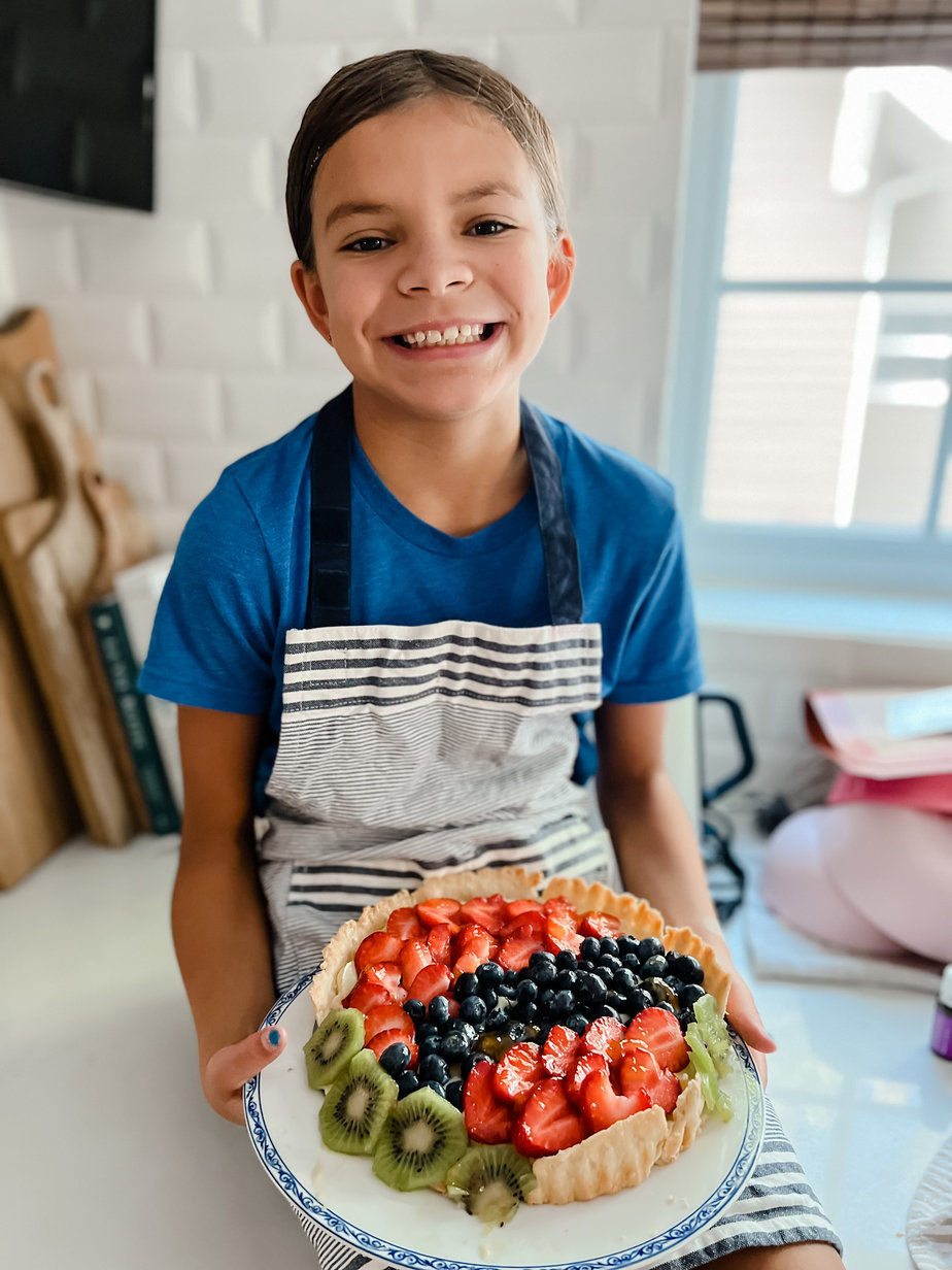 Little girl sitting on a counter holding a french fruit tart and wearing blue t-shirt and striped apron