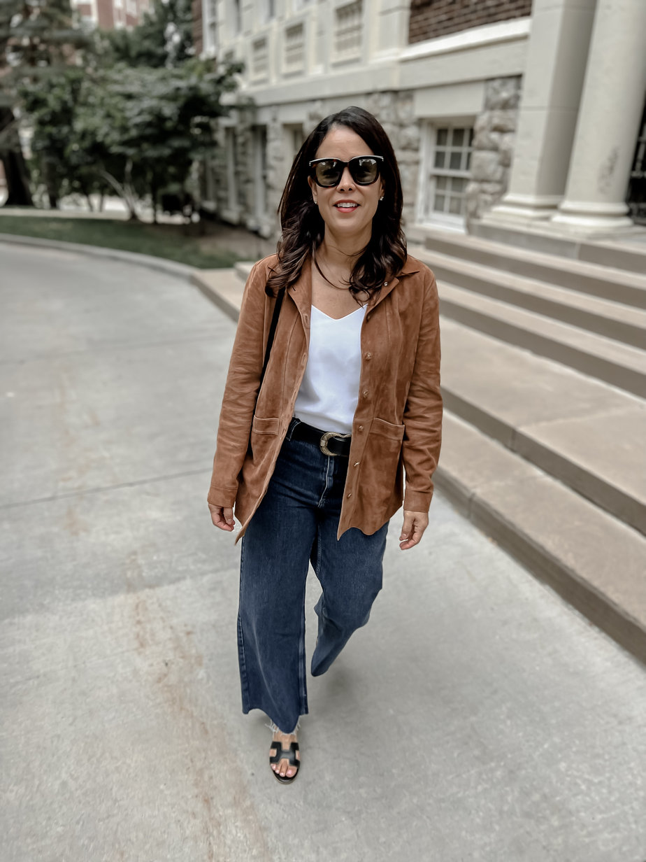 Wearing suede fall transition jacket, white tank top, denim, black sandals, and sunglasses walking down the street with sunglasses.