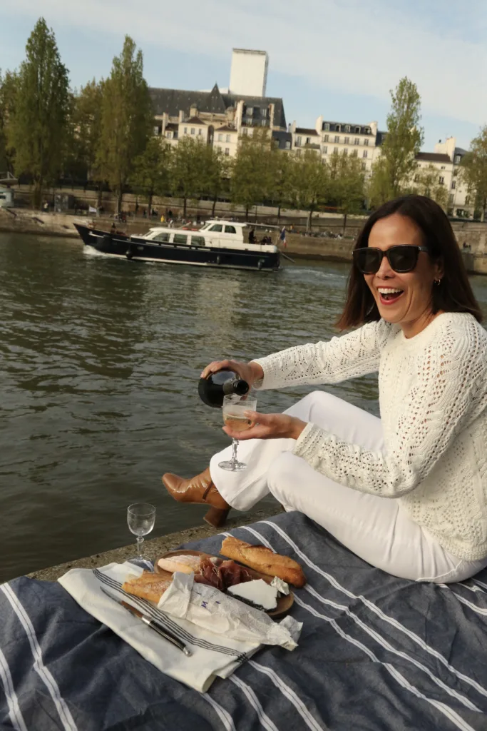 Having a picnic in Paris on the Seine, wearing a white sweater and white jeans, pouring glass of champagne.