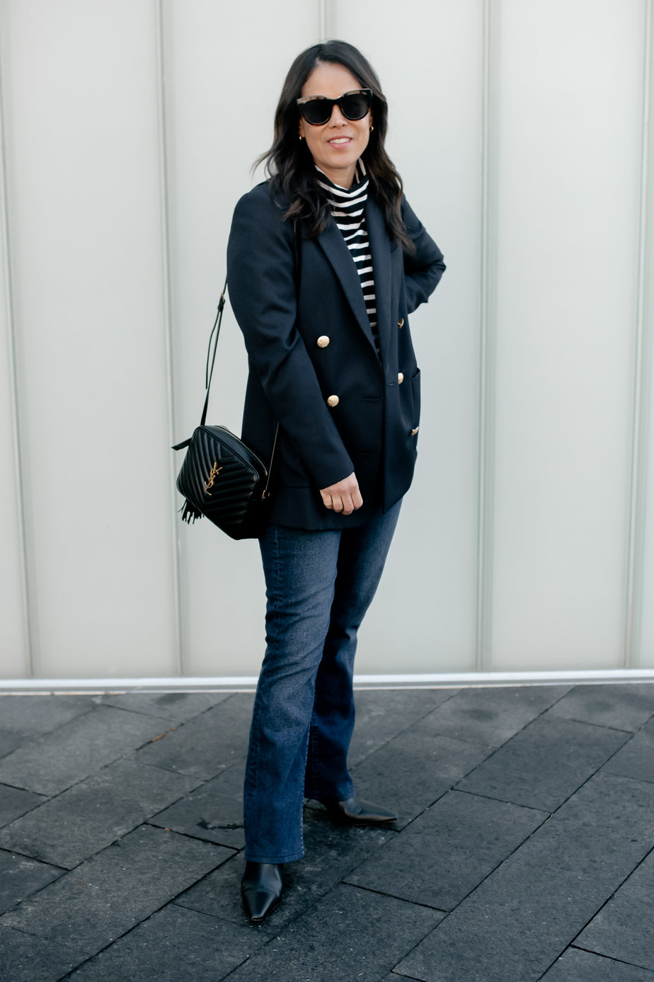 Lady wearing navy blazer with gold buttons and dark denim and boots standing against a grey wall.