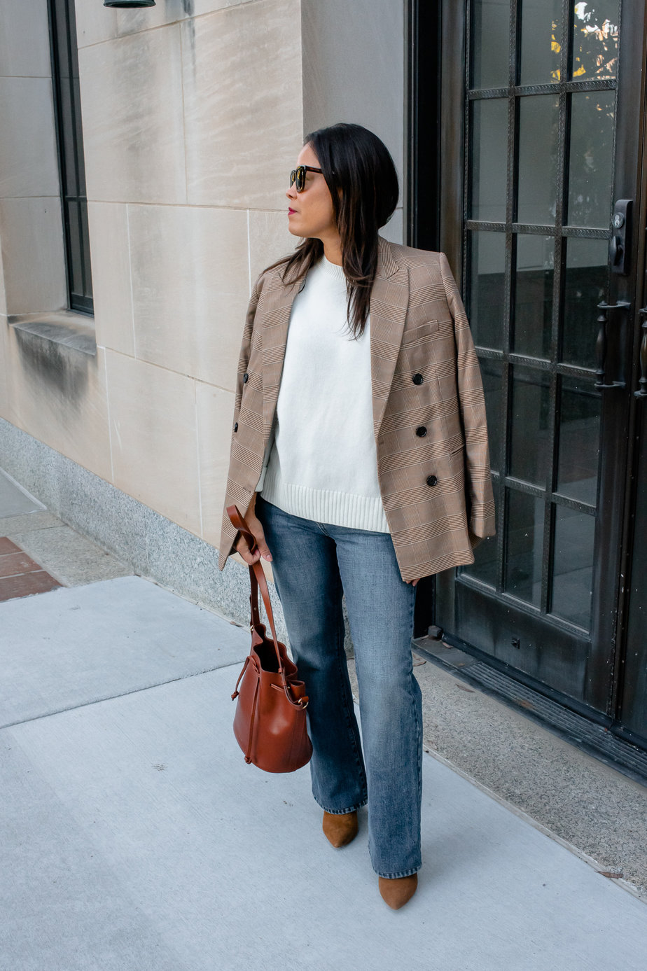 girl standing in front of black doors wearing white sweater, jeans, plaid blazer