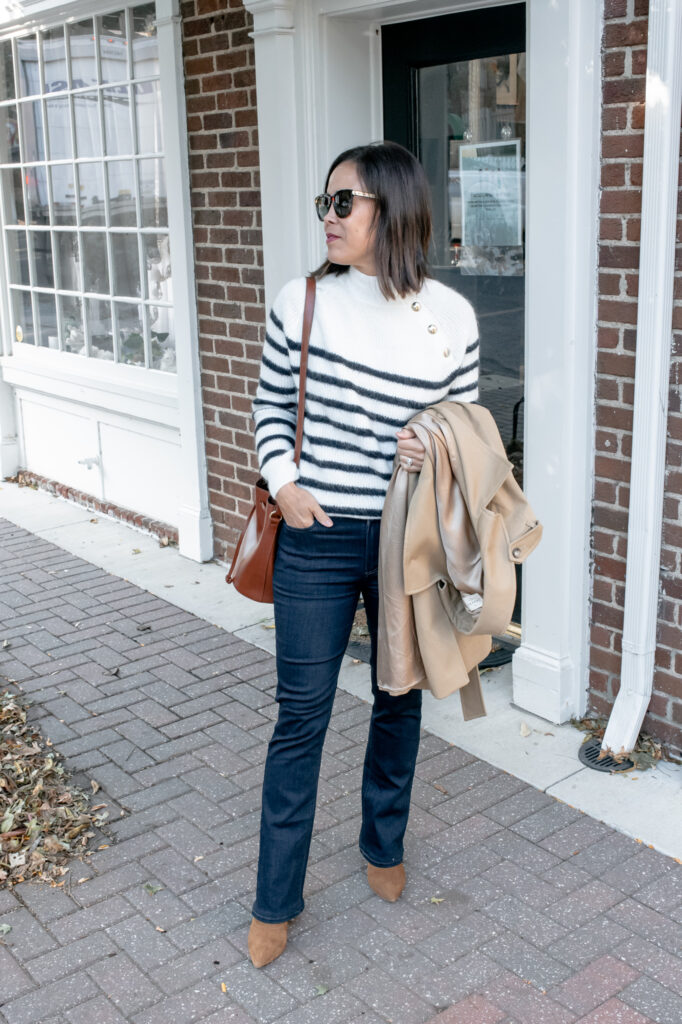 Lady wearing striped sweater with dark blue jeans, trench coat, and boot standing against a wall