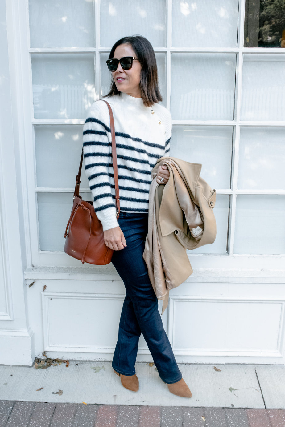 Lady standing against a wall wearing striped sweater and holding a trench coat with brown leather bag