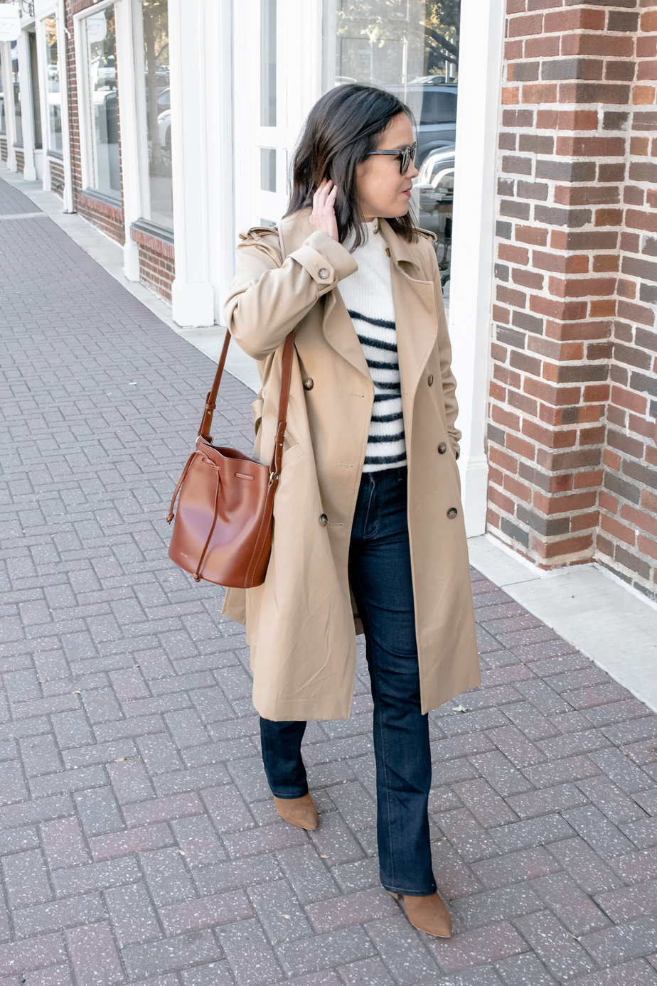 Wearing a trench coat with striped sweater, dark denim, brown boots, walking down the street part of fall capsule wardrobe for 2022.