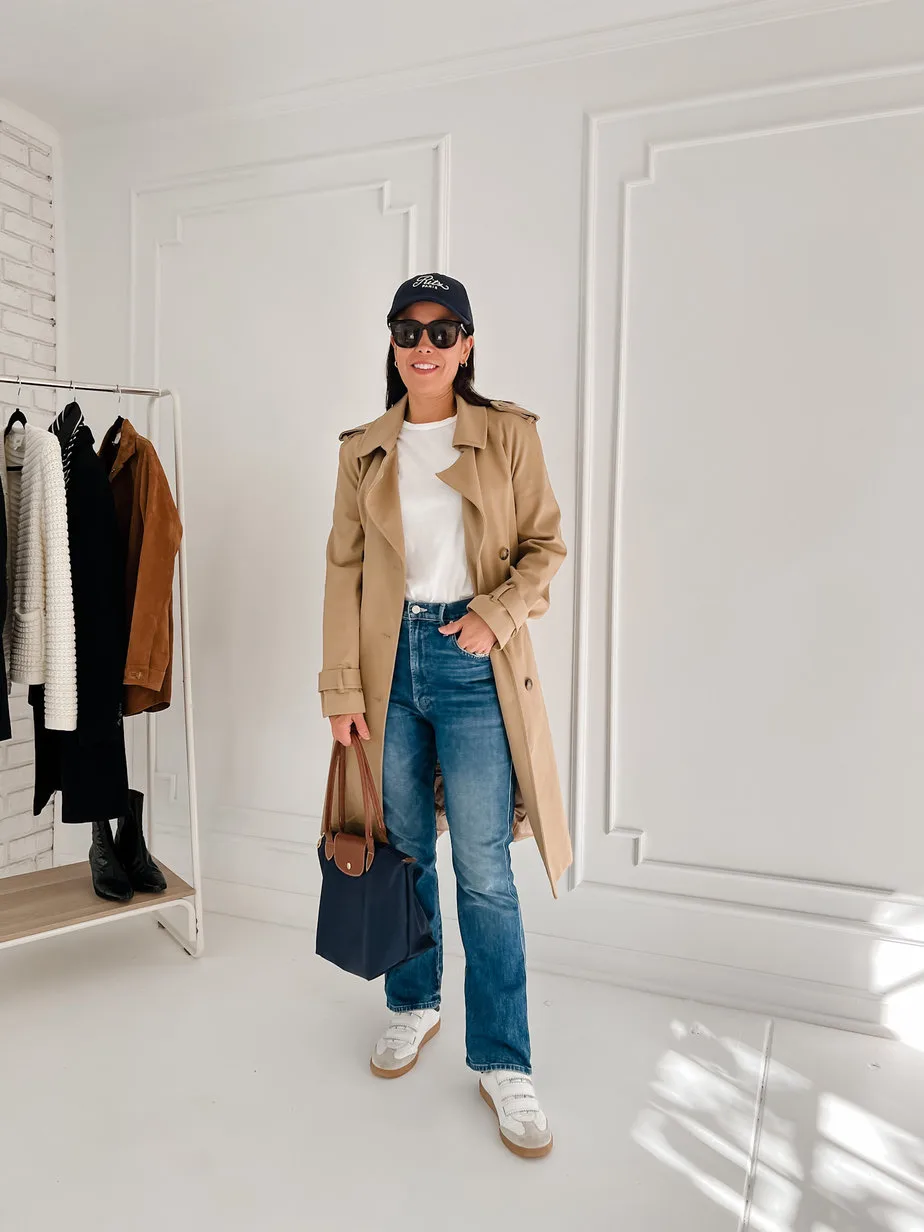 Woman wearing trench coat with white tee, blue denim, white sneakers, navy hat standing in front of a white wall.