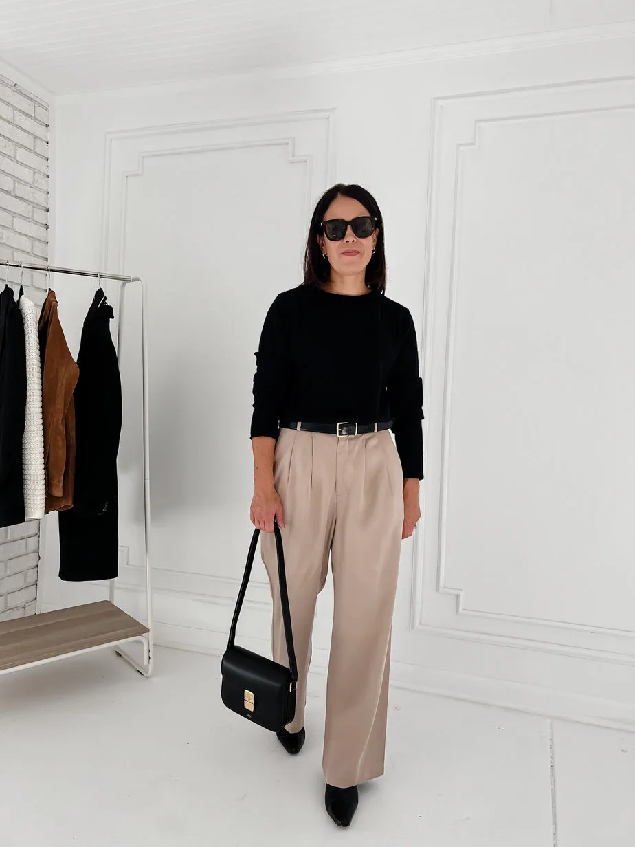 Wearing black sweater, brown trousers, boots, black handbag against black wall.