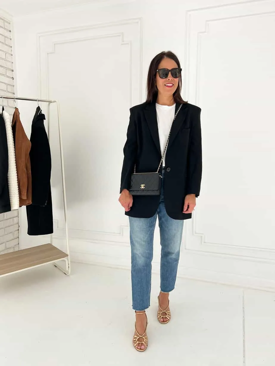 Wearing black blazer with denim, loafers, standing against white wall.
