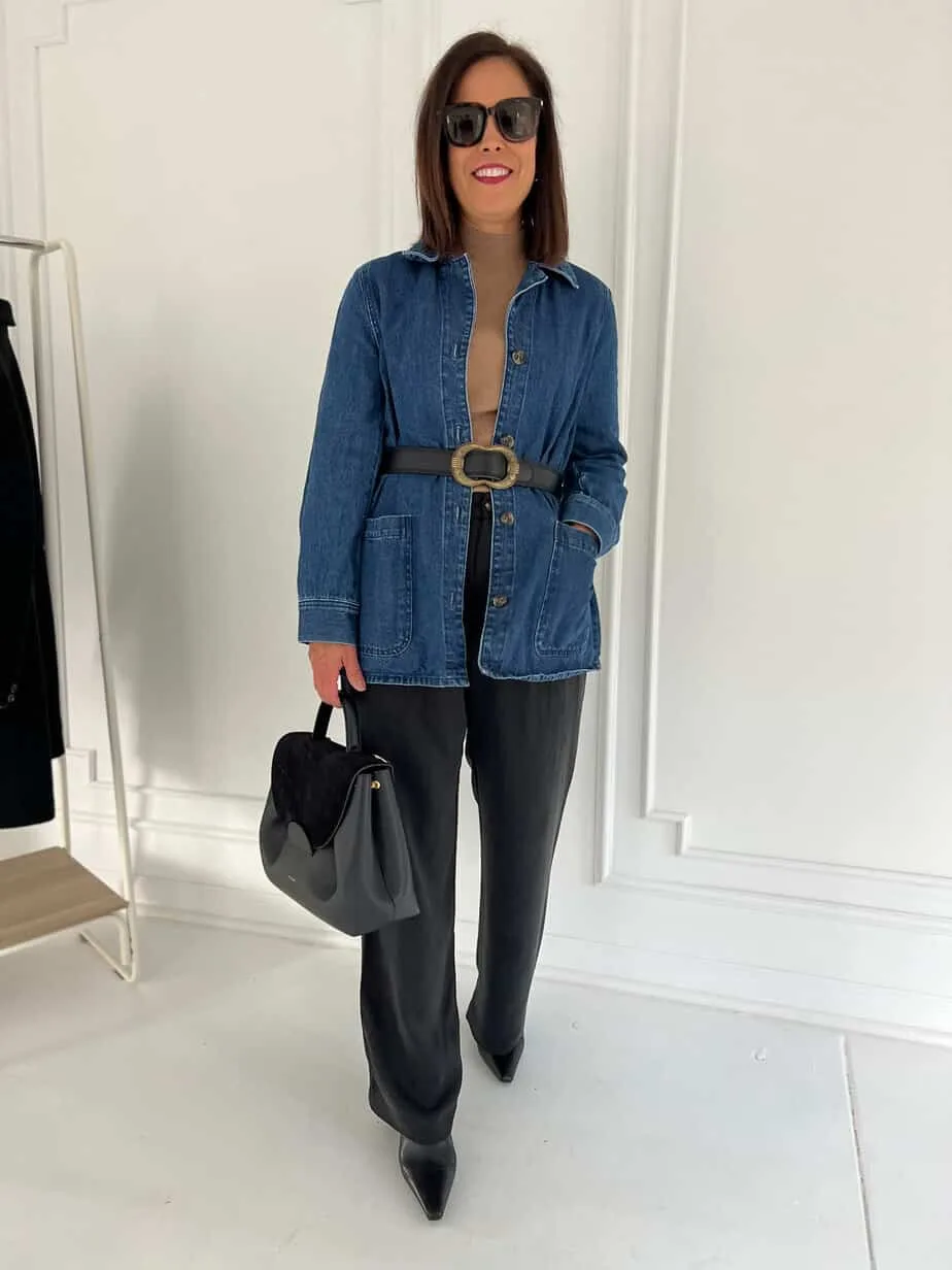 wearing denim jacket with black pants, turtlneck and black bag against a white wall.