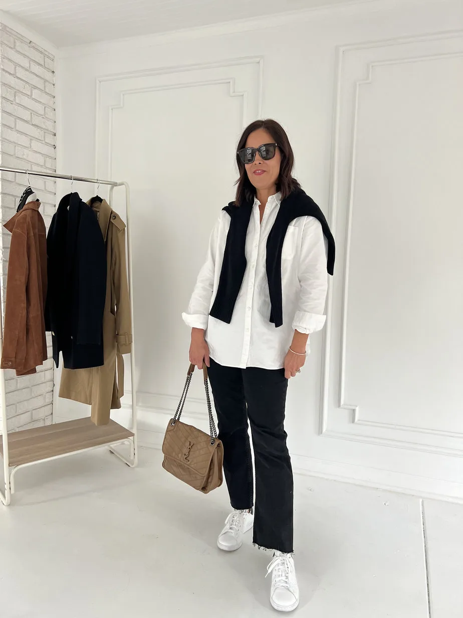 Lady wearing white button down with black denim and sneakers. Has a black sweater over her shoulder and carrying brown leather bag.