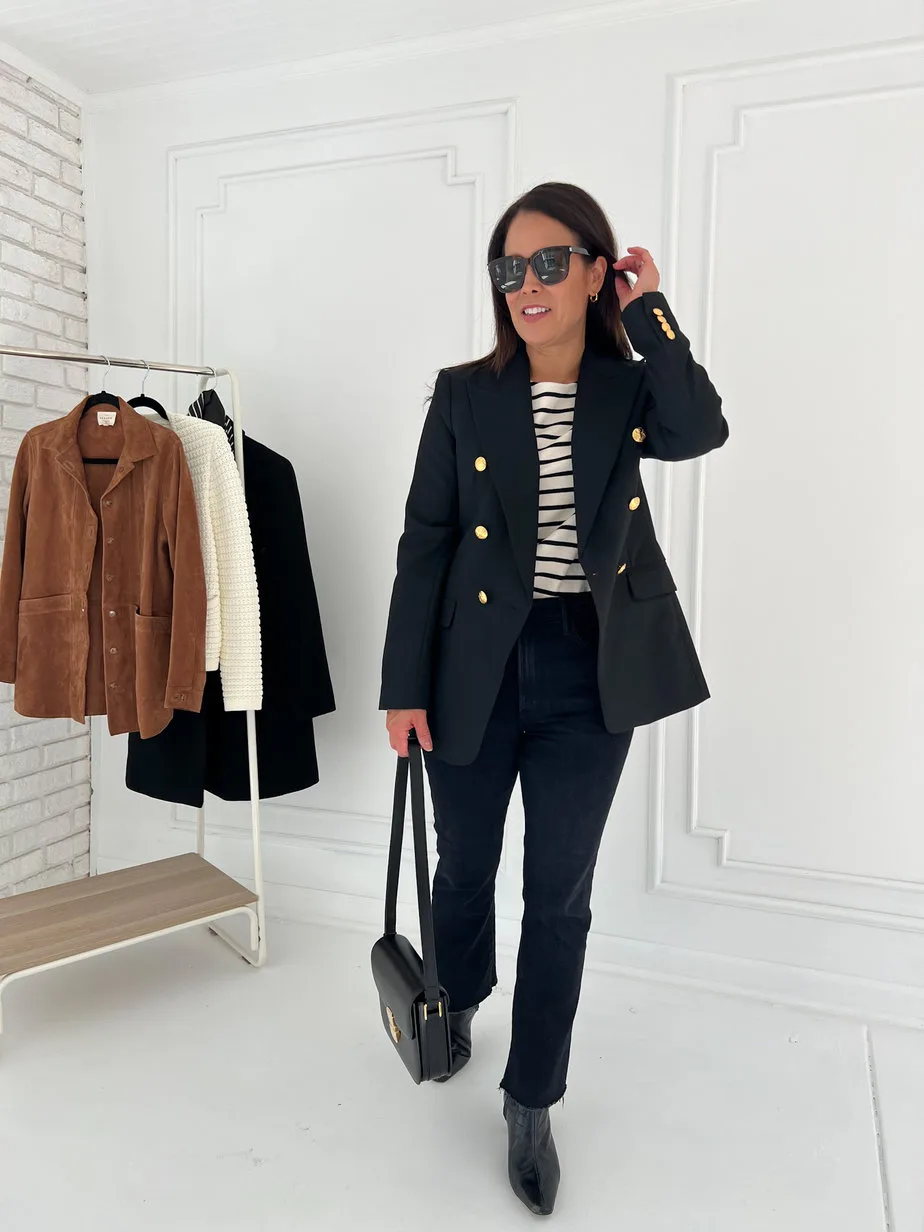 Woman wearing navy blazer, striped top, black denim, carrying a black handbag standing in front of a white wall for a french daily look