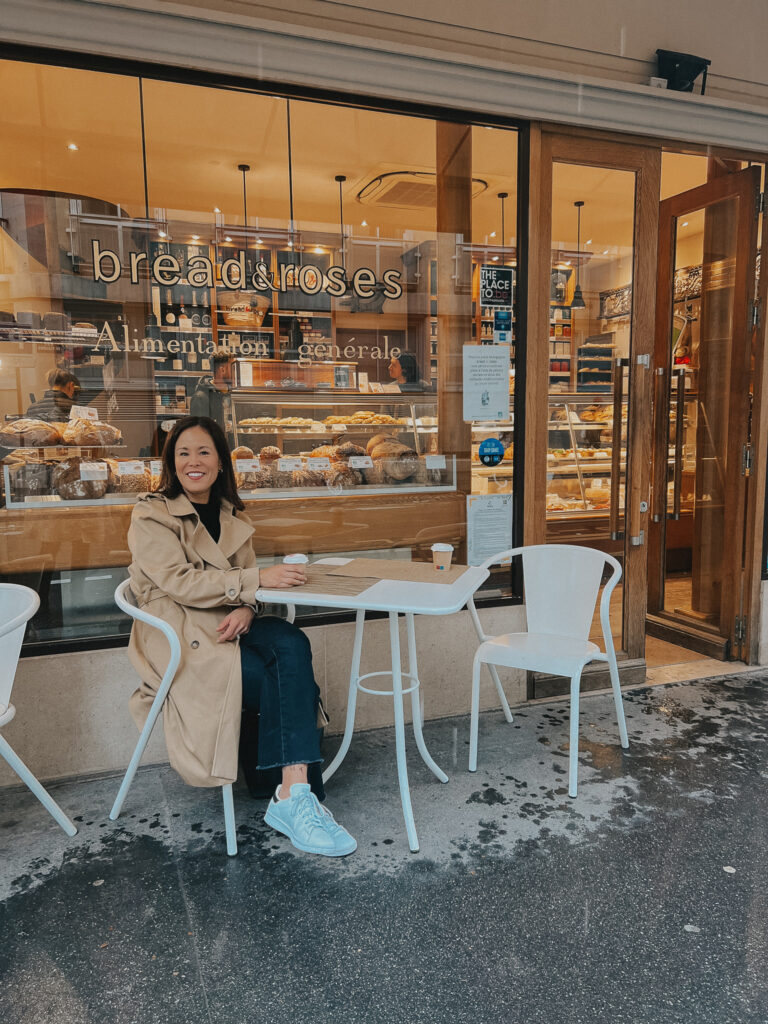Wearing trench coat with black sweater, jeans, and sneakers sitting at a cafe in Paris.