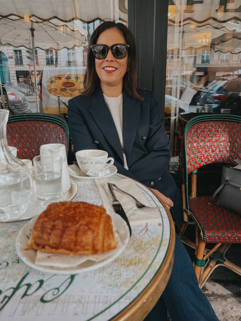 Sitting at a cafe in Paris with a croissant wearing navy blazer and cream sweater.