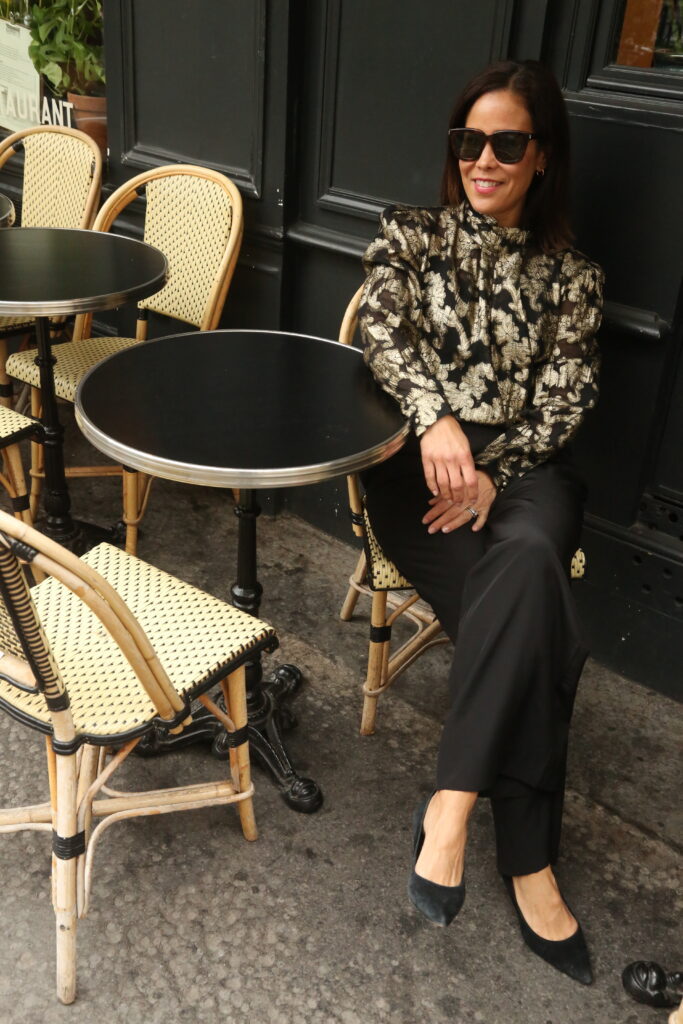Woman sitting at a cafe wearing black silk pants and black and gold top with pumps for a classic and chic holiday look.