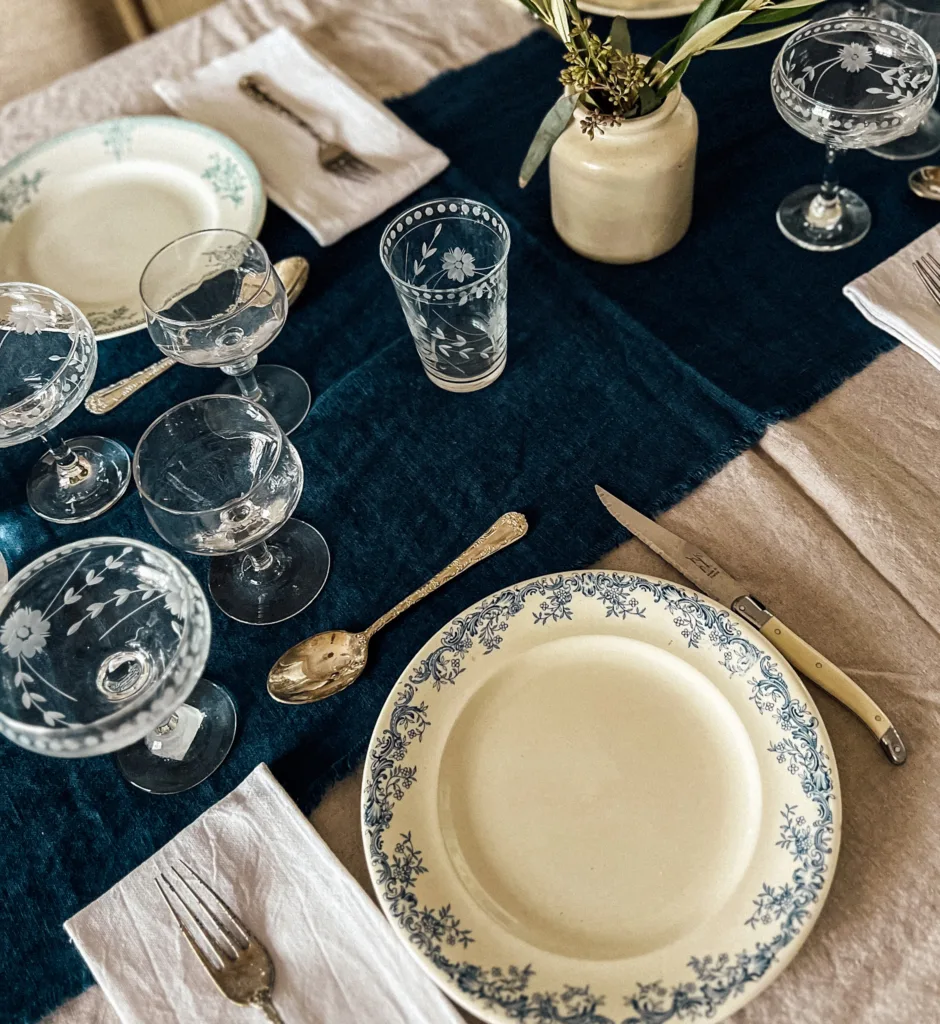 Antique plates, glassware, silverware and napkins on a blue table runner for a French dinner party.