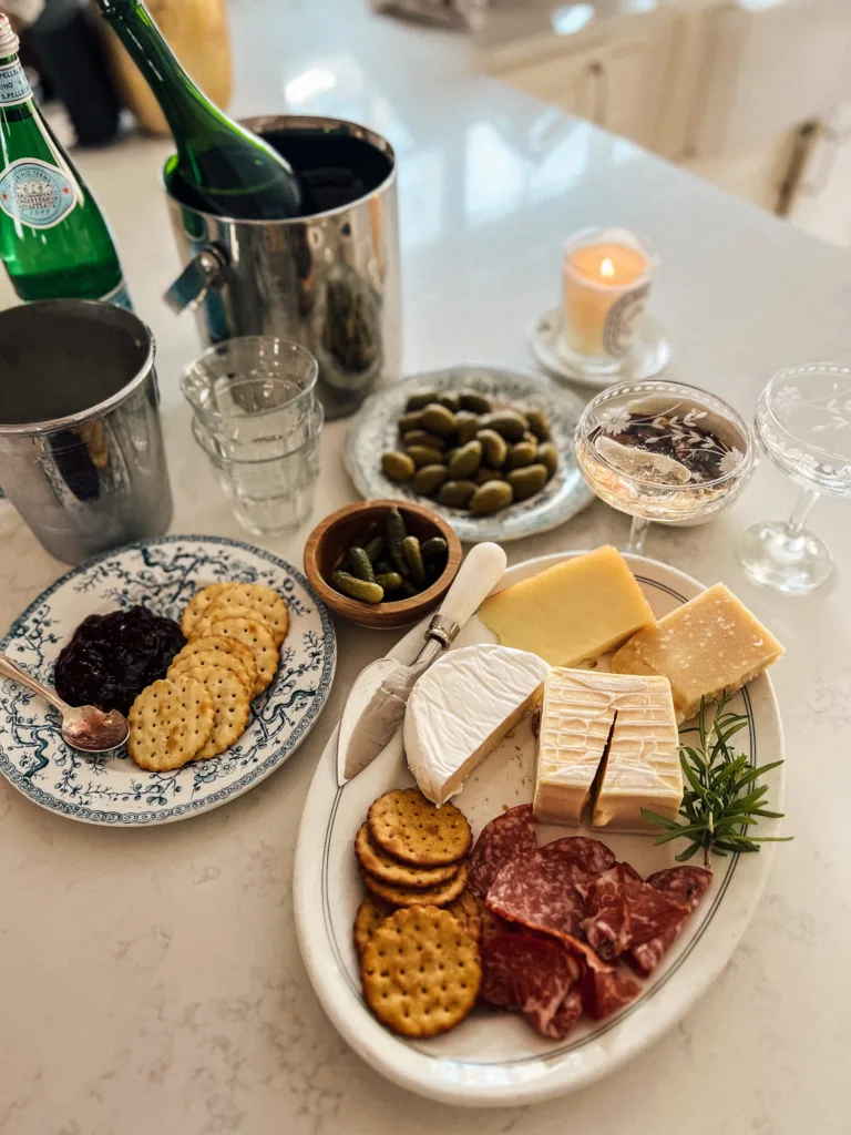 cheese platter sitting on a marble countertop with olives, champagne bucket, and crackers and jam.