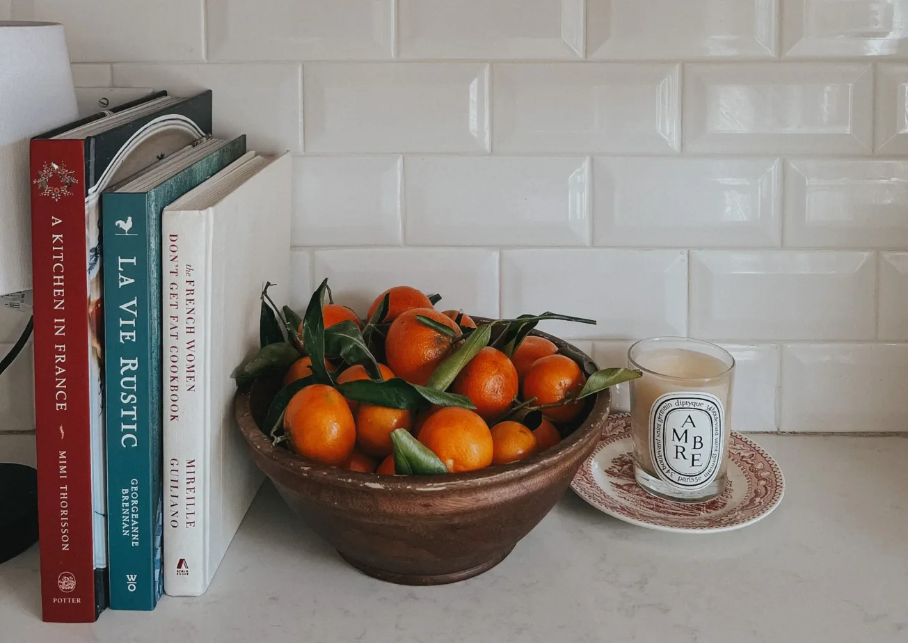Bowl of oranges in wood bowl with candle and cookbooks next to it.