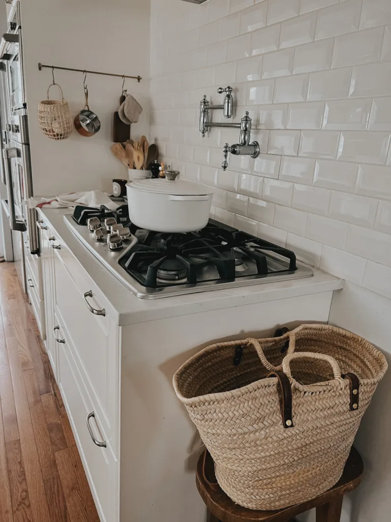 Stovetop with White Le Crueset Dutch Oven, pot filler, market bag on a brown stool.